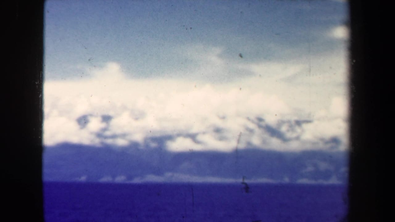 1944 Papua New Guinea aerial landscape showing open water, distant forested mountains, and bright clouds with blue-purple film shift.