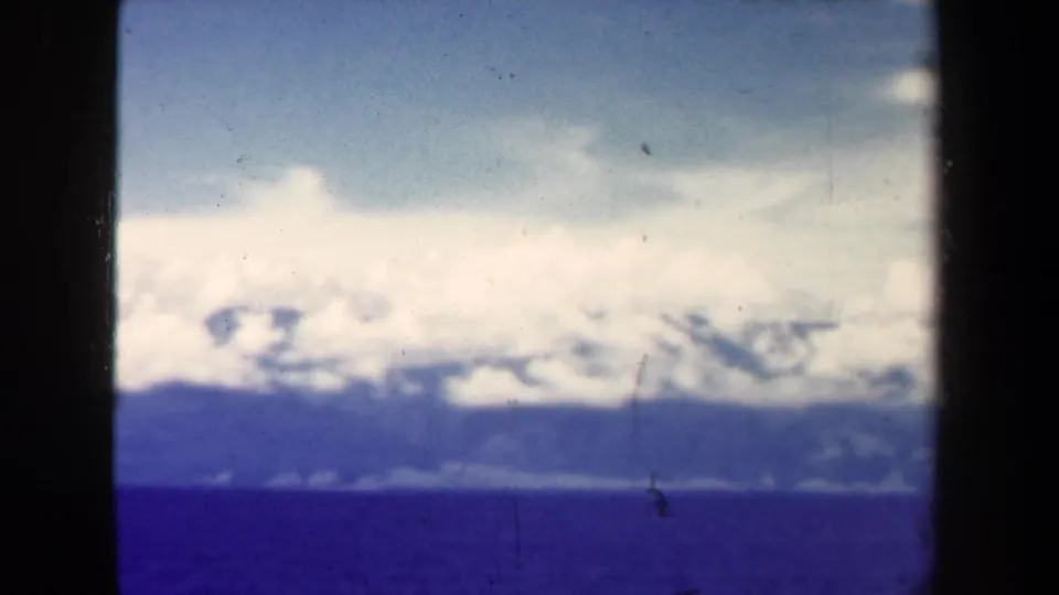 1944 Papua New Guinea aerial landscape showing open water, distant forested mountains, and bright clouds with blue-purple film shift.