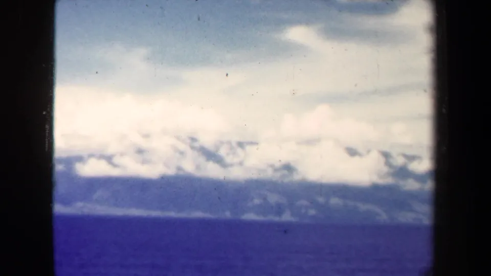 1944 Papua New Guinea 8mm frame showing blue water, a distant mountain range, and layered clouds from a moving aerial viewpoint.