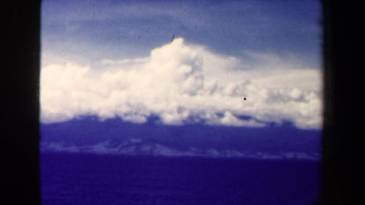Large white cumulus clouds over dark blue sea with a hazy shoreline in the distance.