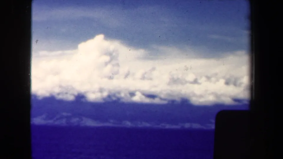 1940s shipboard 8mm view near Papua New Guinea with clouds, ocean, distant land, and a dark foreground obstruction.