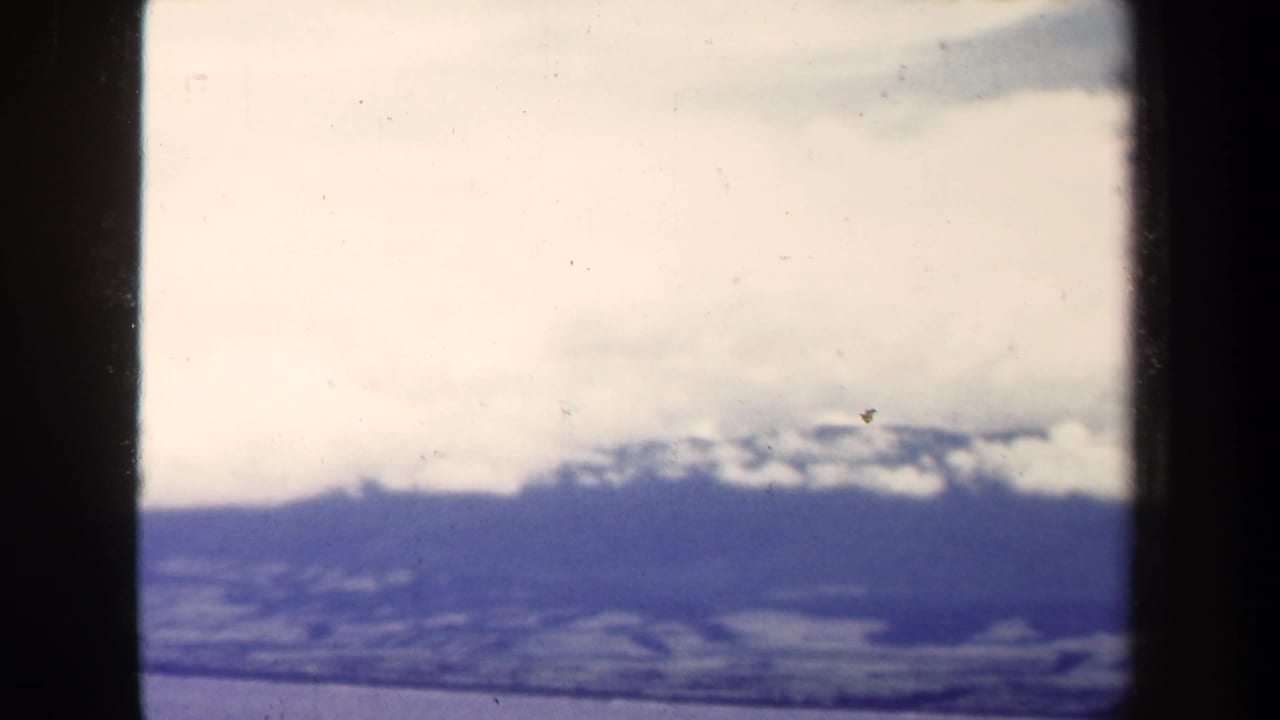 1944 8mm Papua New Guinea view of distant snowy mountain ridges, low clouds, and ocean below, with dark film-edge shadows.