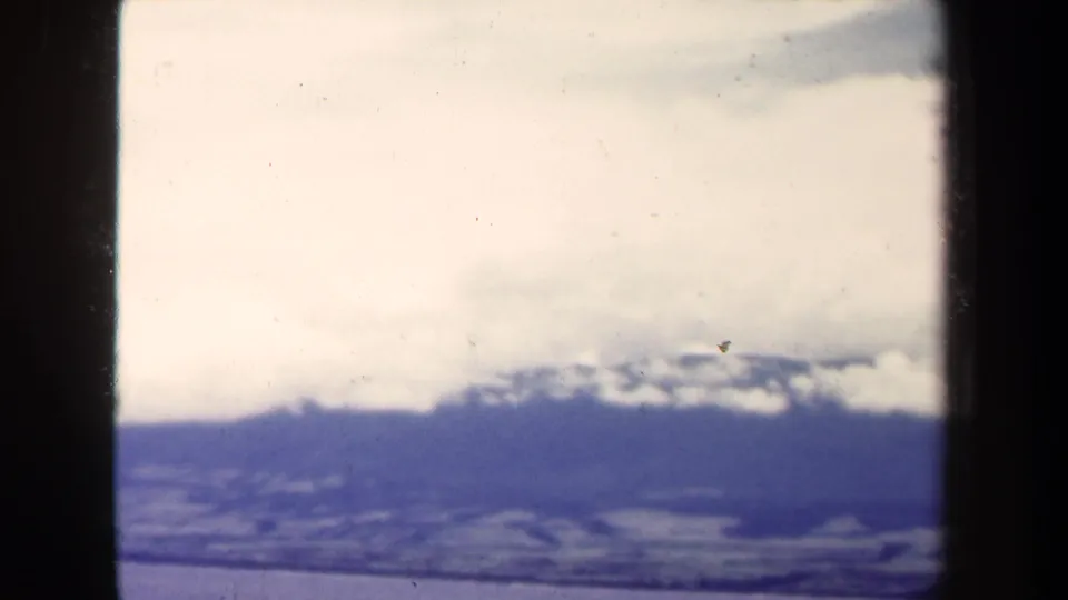 1944 8mm Papua New Guinea view of distant snowy mountain ridges, low clouds, and ocean below, with dark film-edge shadows.