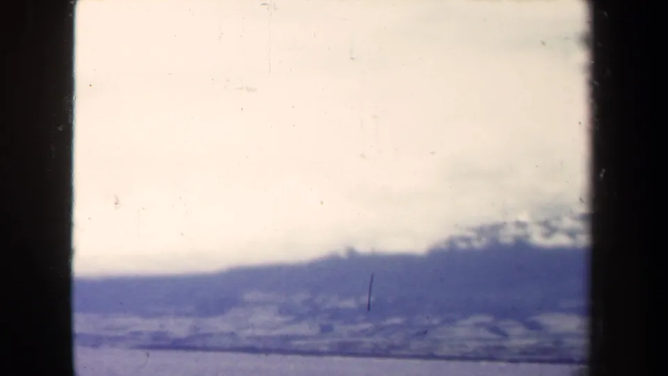 Faded 1944 8mm frame showing distant Papua New Guinea mountains, cloud cover, and sea beneath a bright sky.