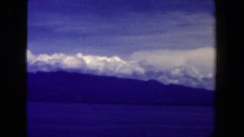 Blue-shifted 1940s 8mm view of snow-capped Papua New Guinea mountains beyond dark ocean water and layered clouds.