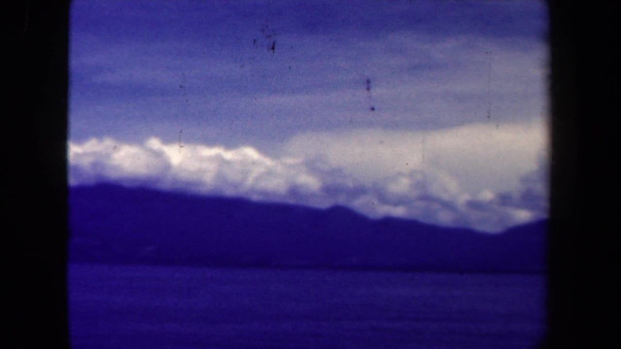 1944 8mm Papua New Guinea landscape showing a lake, dark mountain ridge, and layered clouds under a blue-purple sky.