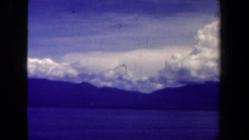 1944 8mm Papua New Guinea landscape with a low mountain ridge, lake water, bright cloud bank, and saturated blue-purple color.