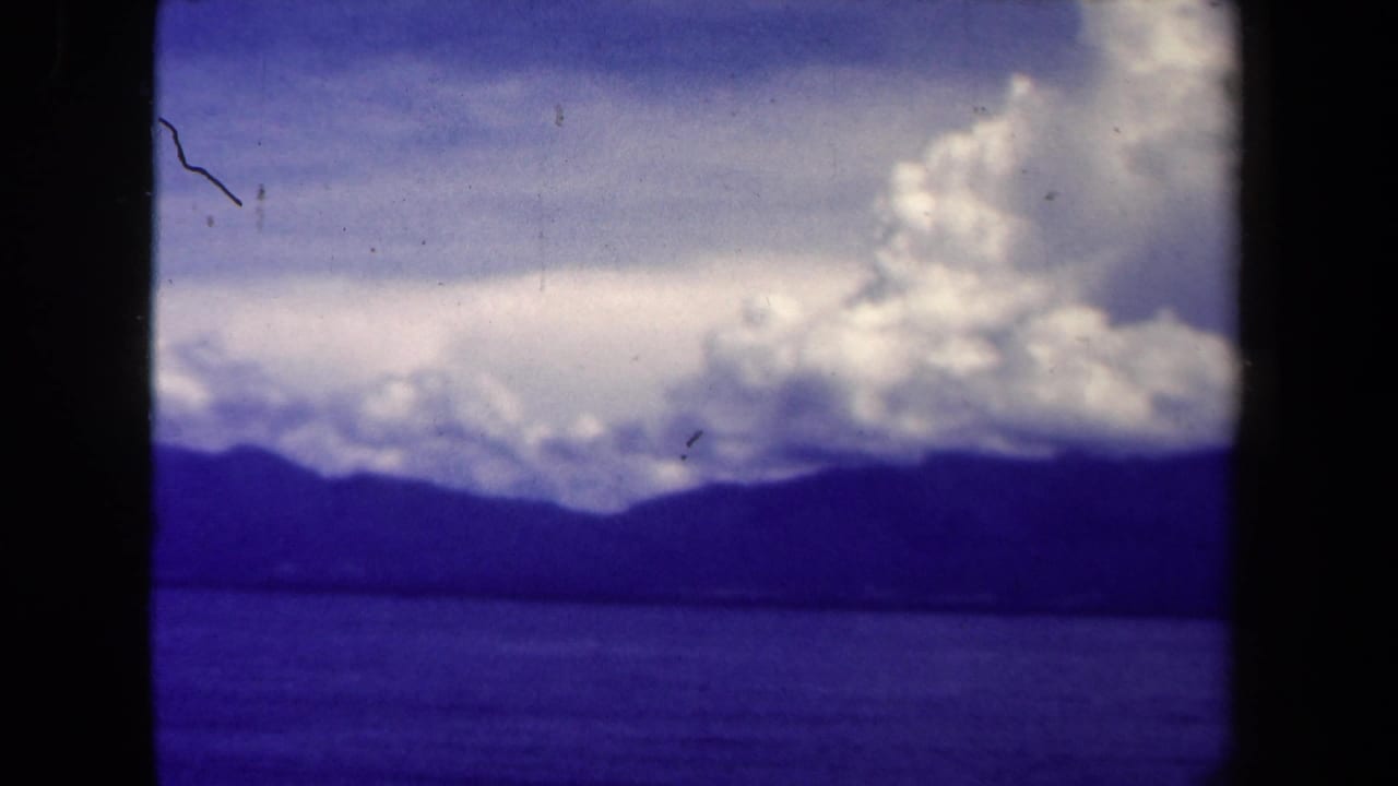 1944 Papua New Guinea 8mm frame showing water, a dark mountain ridge, and bright clouds under a blue sky.