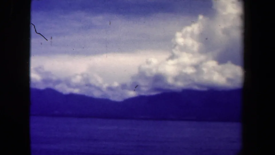 1944 Papua New Guinea 8mm frame showing water, a dark mountain ridge, and bright clouds under a blue sky.