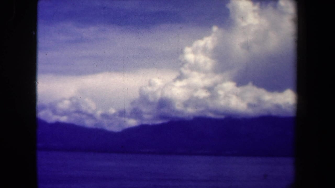 1944 Papua New Guinea landscape frame with a low water horizon, dark mountains, and thick white clouds drifting overhead.
