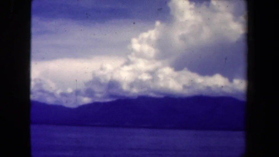 1944 Papua New Guinea 8mm still with blue water, a silhouetted mountain range, and a luminous cloud mass above.