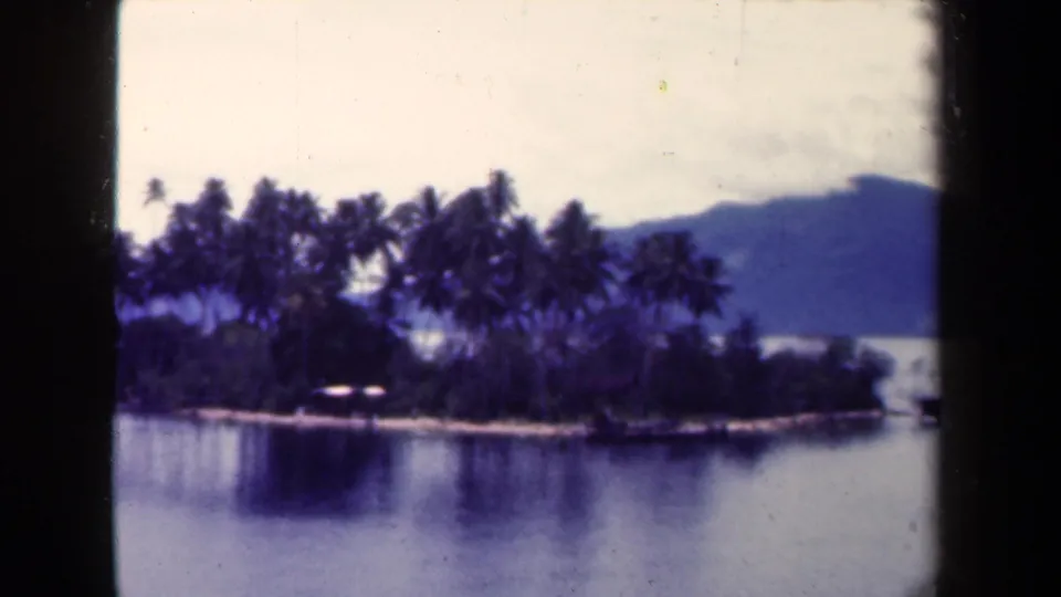Vintage 8mm frame of a tropical island shoreline in Papua New Guinea, with palms, reflections, and a blue mountain ridge.