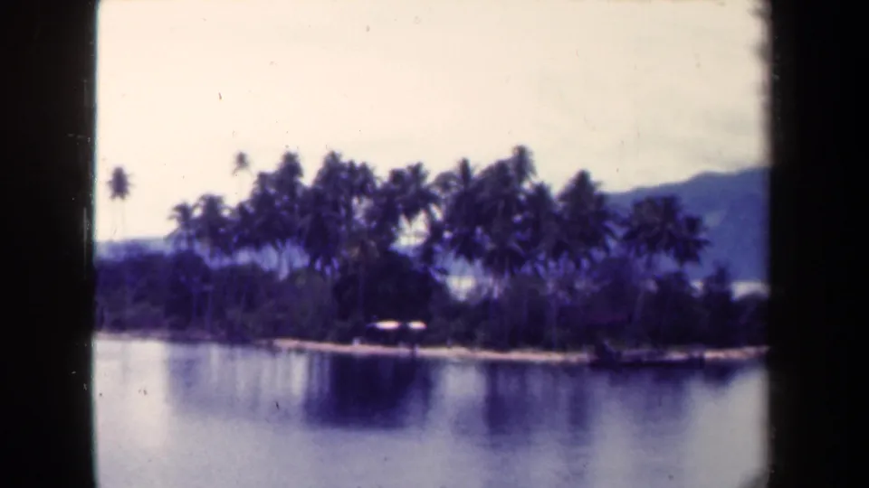 1944 Papua New Guinea 8mm frame showing calm water, a small palm-lined landmass, a low structure, and hazy hills beyond.