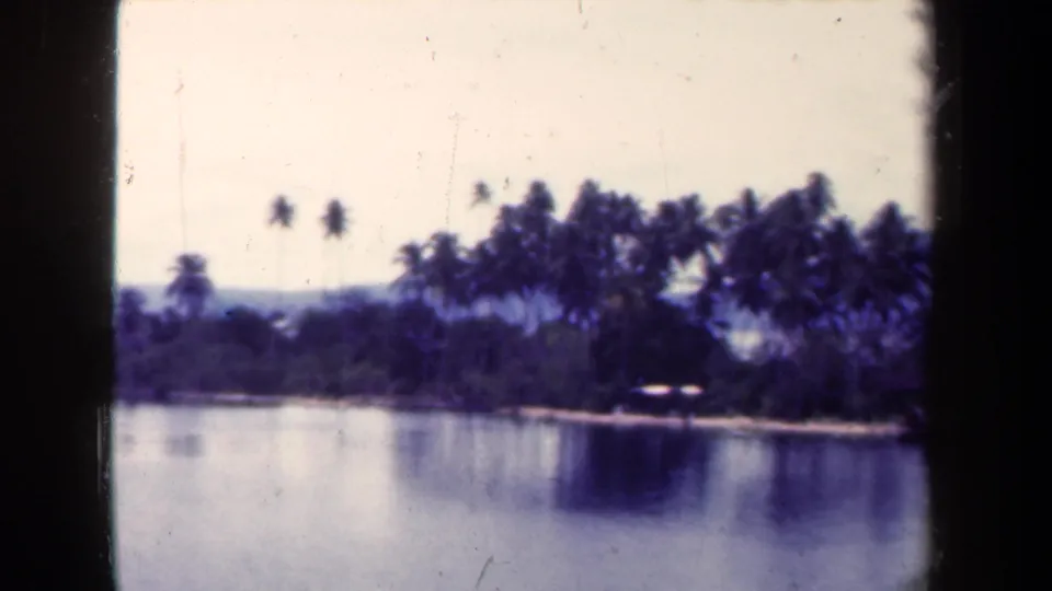 1944 Papua New Guinea 8mm frame showing a slow view of palms, a small shoreline structure, calm water, and hazy background hills.