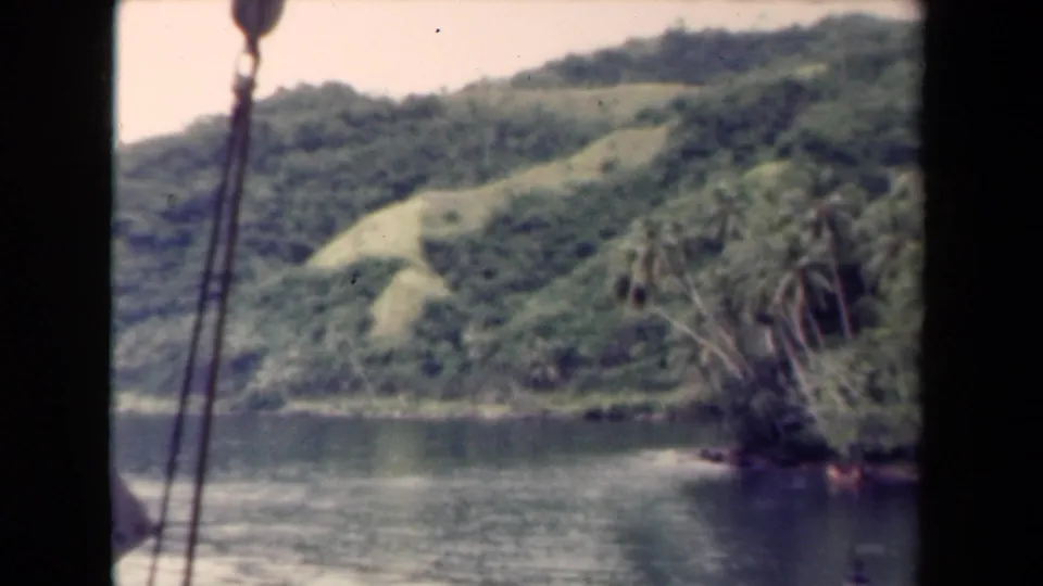 1944 Papua New Guinea home-movie frame with boat rigging in the foreground and a wooded hillside beyond the water.
