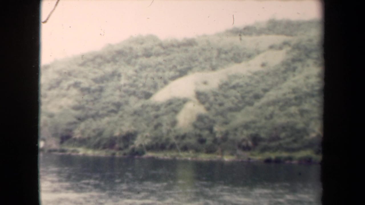 1944 8mm home-movie view in Papua New Guinea of a soft green hillside rising from calm water, with dark film borders visible.