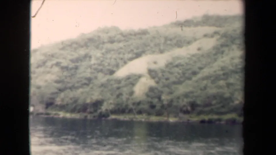 1944 8mm home-movie view in Papua New Guinea of a soft green hillside rising from calm water, with dark film borders visible.