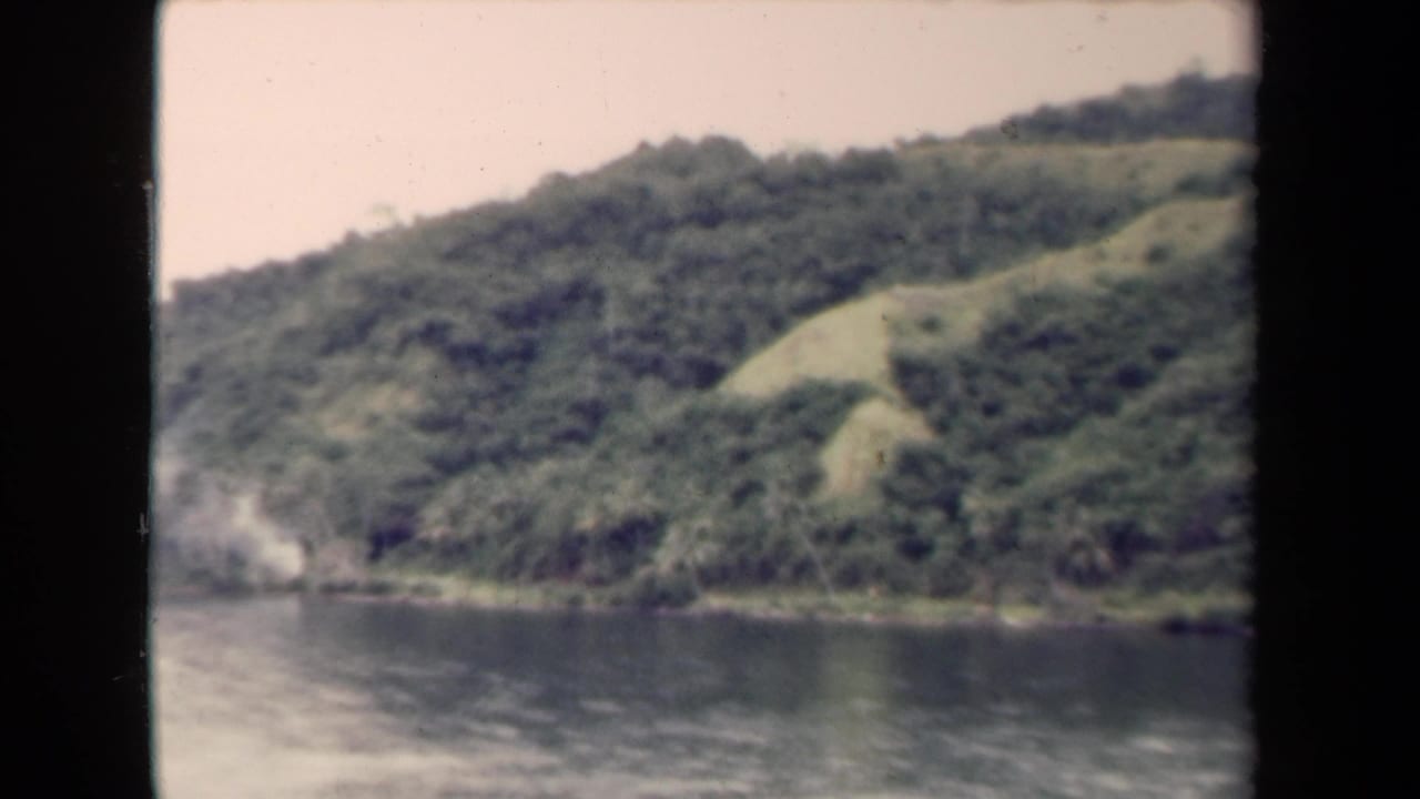 Papua New Guinea landscape frame showing a steep green slope, dense shoreline growth, and reflective water in a muted 8mm scan.