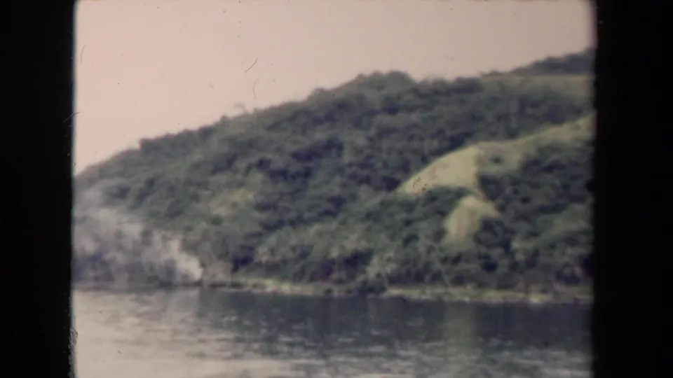 Soft 8mm frame of water below a forested Papua New Guinea hillside, with pale shoreline detail and visible film wear.