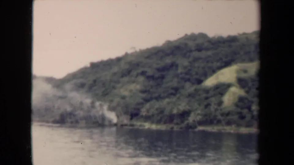 Final 1944 8mm frame looking from water toward a jungle-covered Papua New Guinea hillside under a pale sky.
