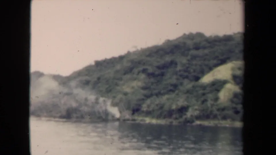 1944 Papua New Guinea 8mm frame showing calm river water below a densely forested hillside and pale sky.