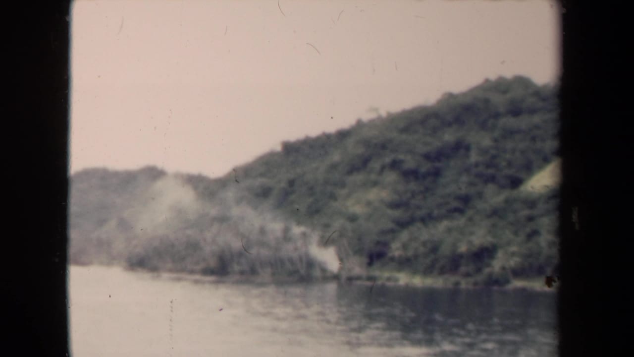 1944 Papua New Guinea river view with green hillside, calm water, and pale smoke or mist along the shoreline.