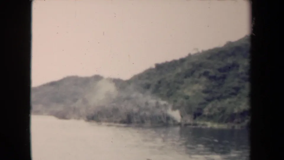 1944 Papua New Guinea 8mm frame showing a broad river, forested slopes, and haze or smoke near the bank.