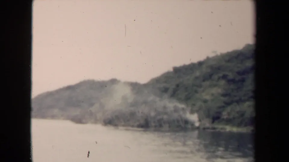 1944 Papua New Guinea river landscape with dense hillside vegetation and a soft smoke or mist plume by the bank.