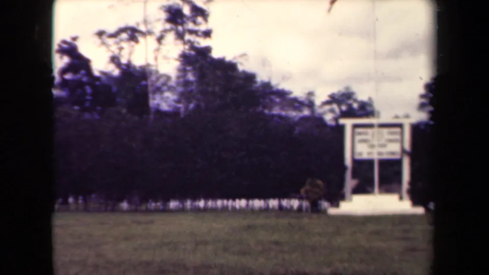1944 Papua New Guinea frame showing a white sign structure set against trees at the edge of an open lawn.