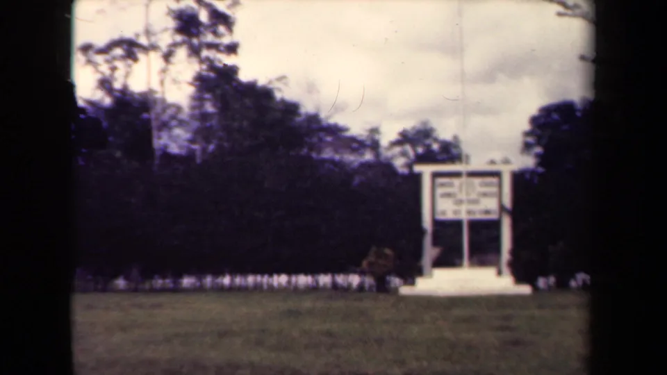 1940s Papua New Guinea 8mm frame showing a white sign structure on a lawn before dark tropical trees.
