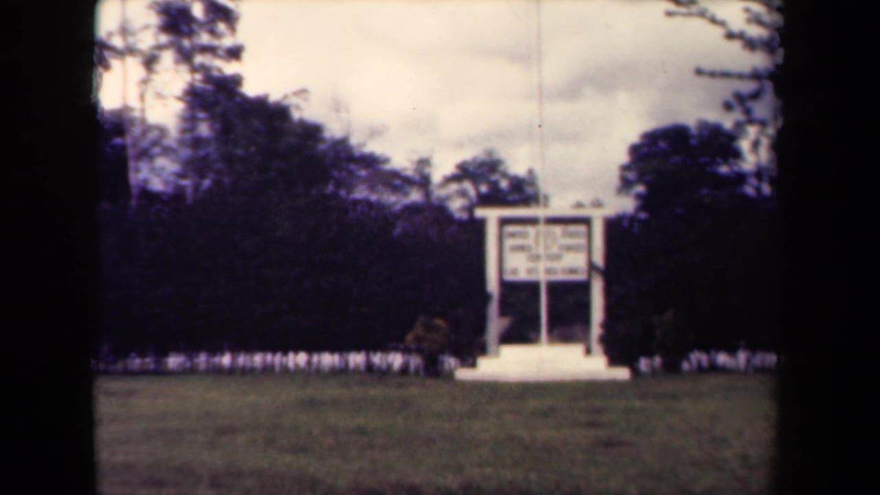 White sign centered beyond a lawn, with trees and an overcast sky in back.