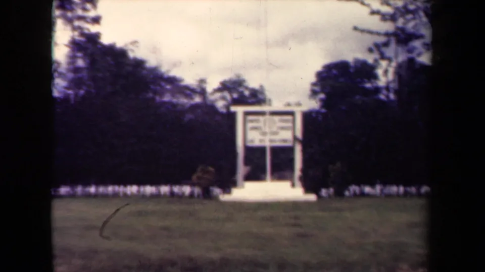 Papua New Guinea 8mm footage shows a white sign with a small shrub beside it, seen from a distance across grass.
