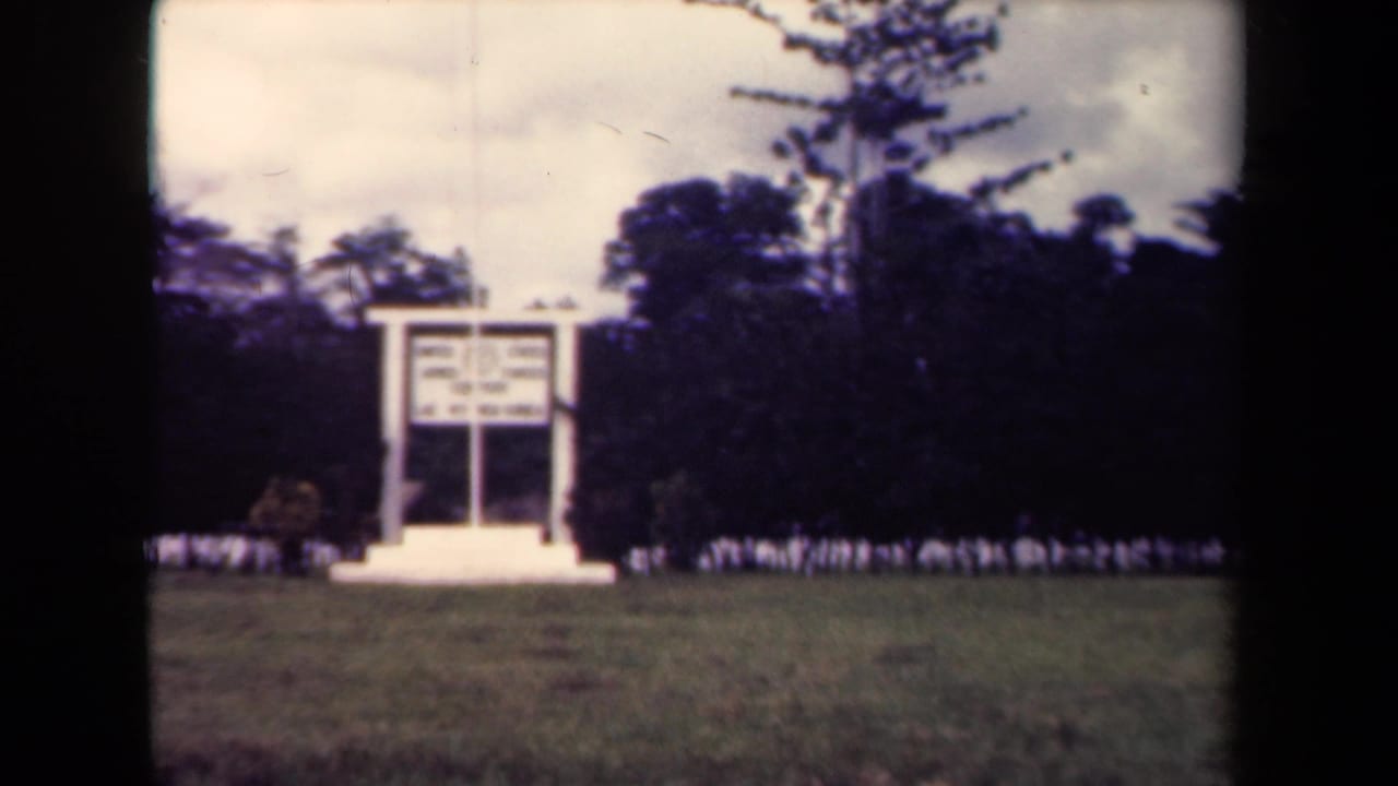 White sign or memorial structure in a grassy field with dense trees behind it.