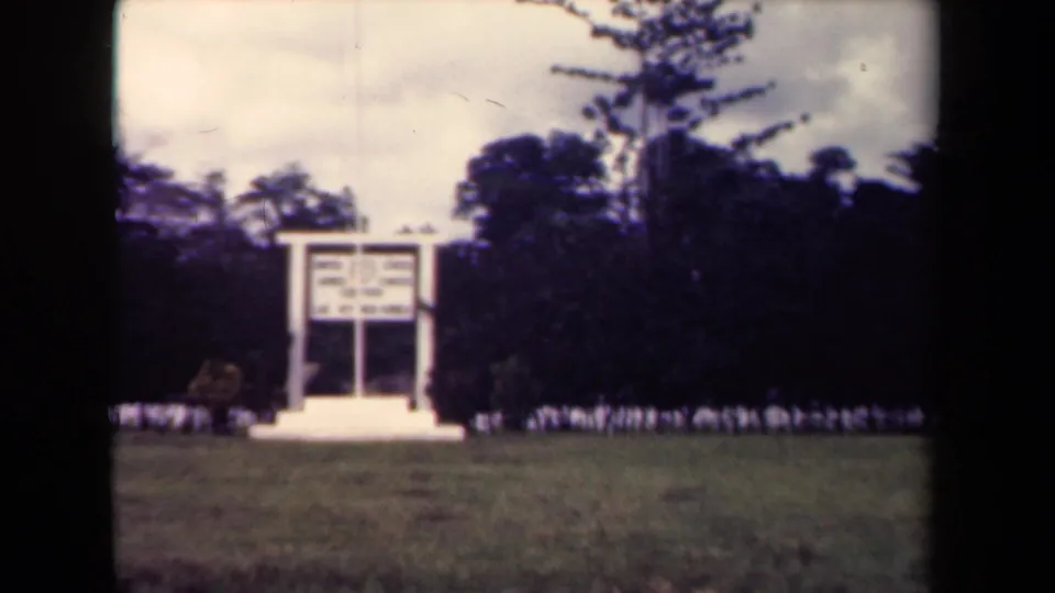 1944 Papua New Guinea 8mm frame showing a white sign or memorial structure on cleared grass with tropical trees and cloudy sky behind.