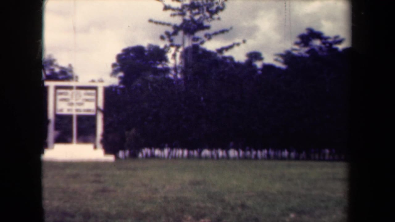 1944 Papua New Guinea frame with a white memorial-like structure, tropical trees, cleared grass, and a distant line of uniformed personnel.