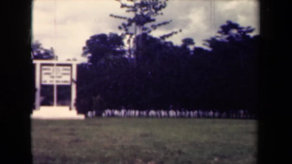 1944 Papua New Guinea frame with a white memorial-like structure, tropical trees, cleared grass, and a distant line of uniformed personnel.