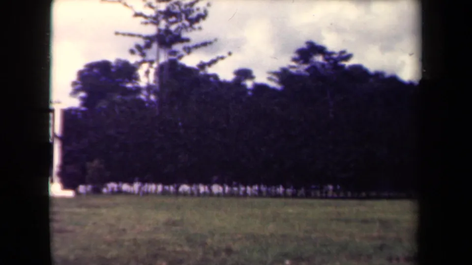 1944 Papua New Guinea 8mm frame during a rightward pan, with the white structure partly cropped and personnel lined along the trees.