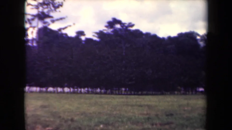 1944 Papua New Guinea frame showing distant uniformed personnel standing along a tropical tree line beyond cleared grass.