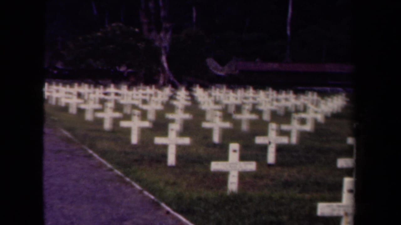 Rows of white crosses fill a grassy U.S. military cemetery in Papua New Guinea, with trees and a path visible beyond the plots.