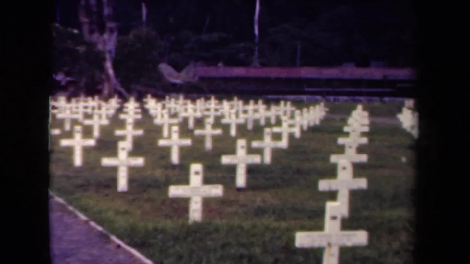 The camera looks across orderly rows of white cross markers in the Lae cemetery, with a path and dense trees framing the graves.
