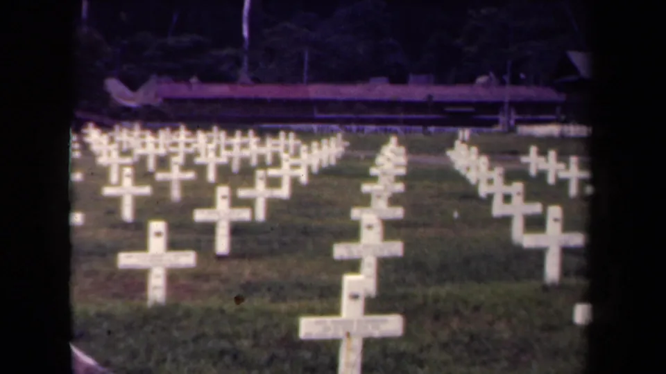 Rows of white military grave crosses extend across the cemetery lawn in 1944 Papua New Guinea footage, with foliage in the background.