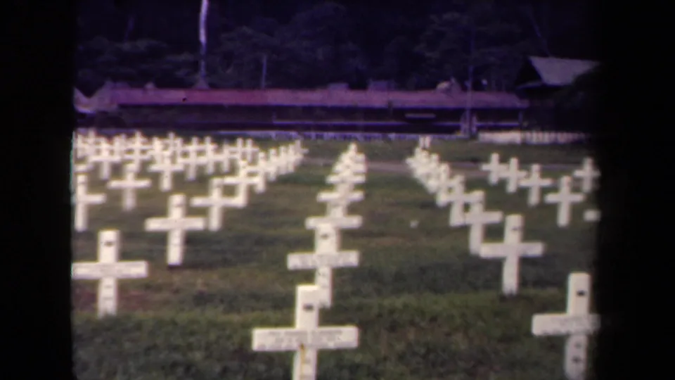 1944 Papua New Guinea 8mm frame showing rows of white grave crosses in a grassy military cemetery with trees and a low background structure.