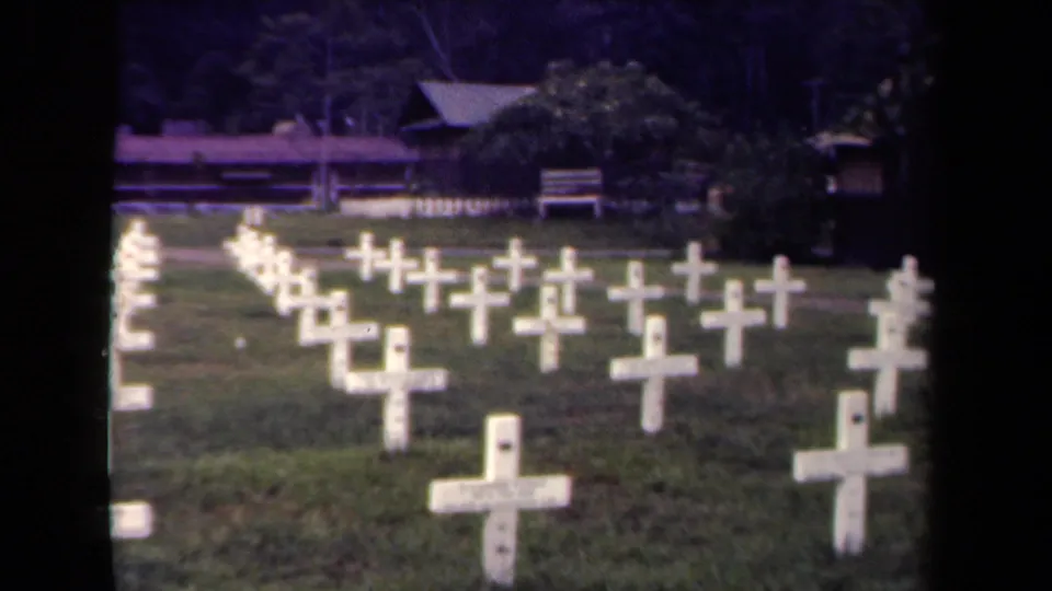 1944 Papua New Guinea 8mm still showing white grave crosses across a lawn with trees and a bench or rail in the background.