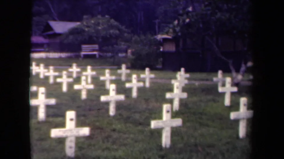 1944 Papua New Guinea frame showing a grassy military cemetery with white crosses, trees, and a shaded structure at the edge of the grounds.