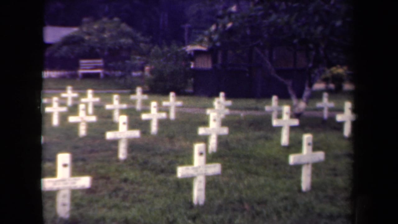 Rows of white cross grave markers on grass with trees and a small structure behind them.