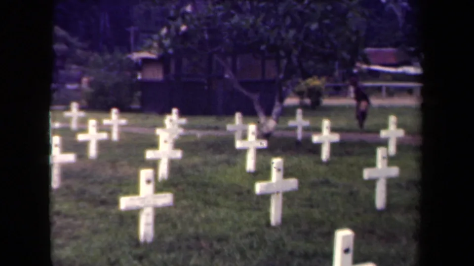 1940s Papua New Guinea cemetery view with rows of white crosses and a distant person walking near the background path.