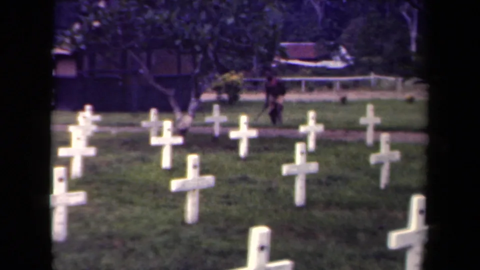 1940s Papua New Guinea home-movie frame looking across cemetery crosses toward a distant walking person and trees.