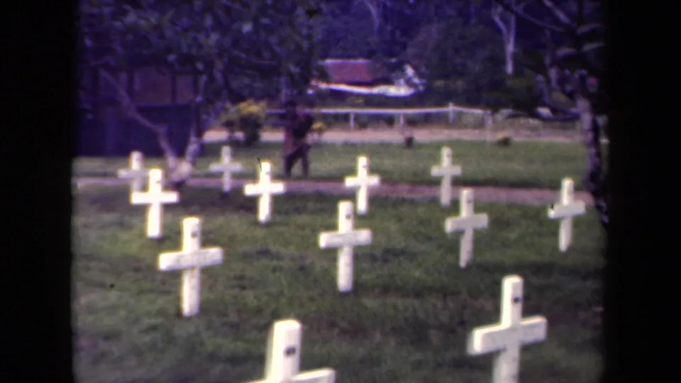 1940s Papua New Guinea cemetery frame showing white cross grave markers, trees, and a distant person near the path.