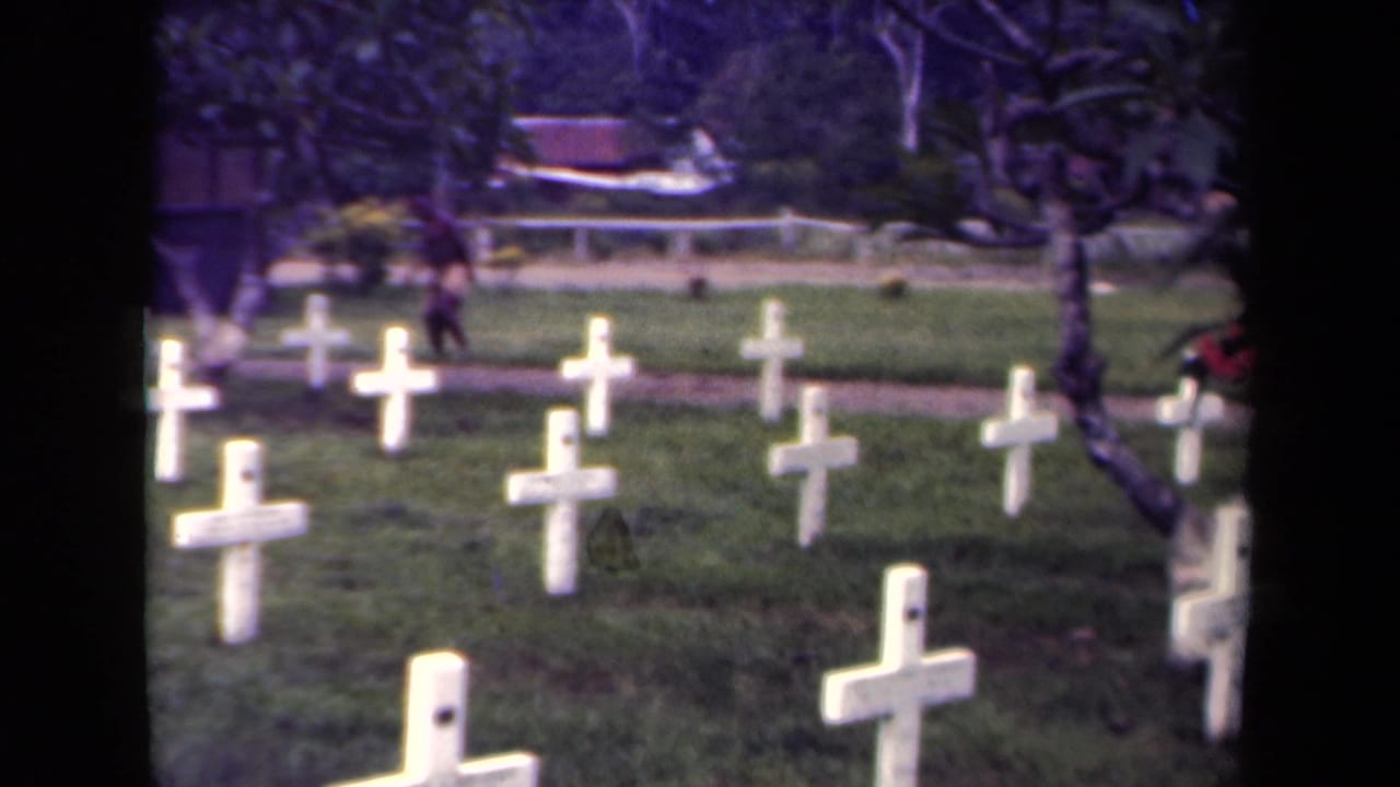 1940s Papua New Guinea 8mm frame showing white wooden cross markers in a green cemetery, with trees and a distant figure near a path.