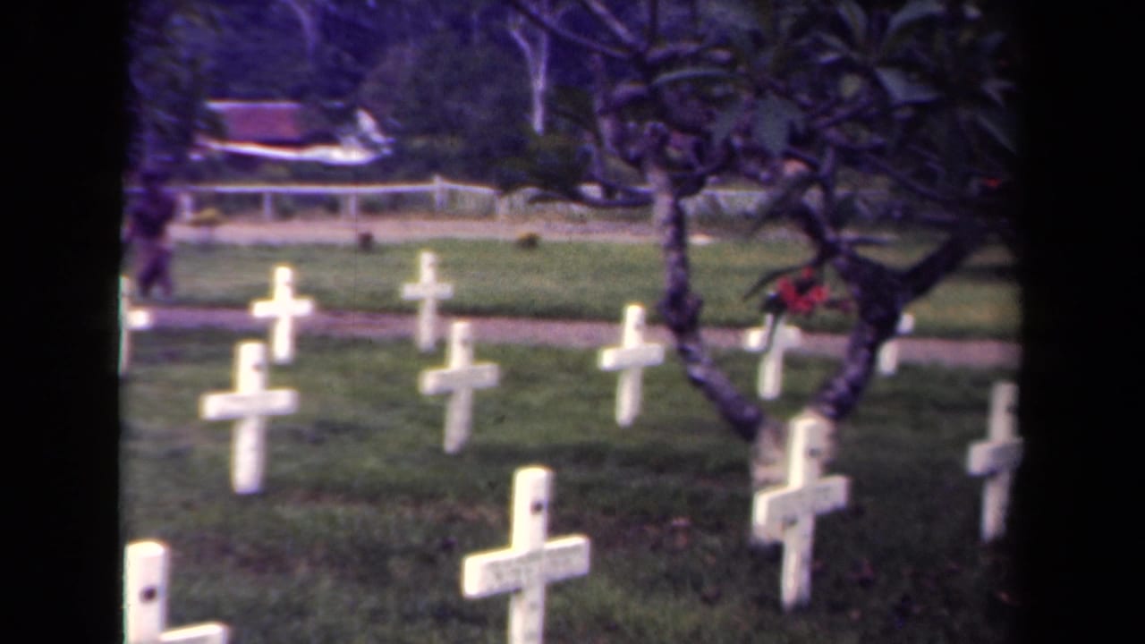1940s Papua New Guinea 8mm frame of white cross grave markers partly framed by a dark tree, with a path and foliage beyond.
